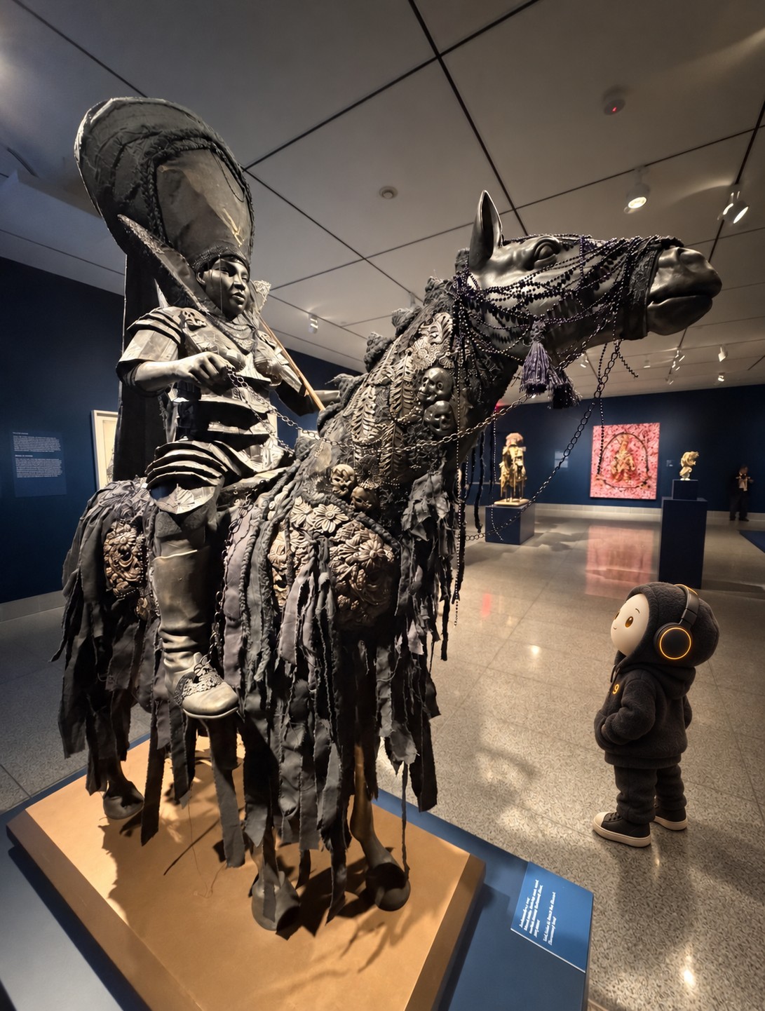 Digital composite of Envoy9, a small ivory-faced figure in a dark hooded jacket and headphones, standing on the right side of an MFAH gallery and looking up at Hew Locke’s large black horse-and-rider sculpture Ambassador 4. The sculpture remains the dominant foreground subject.